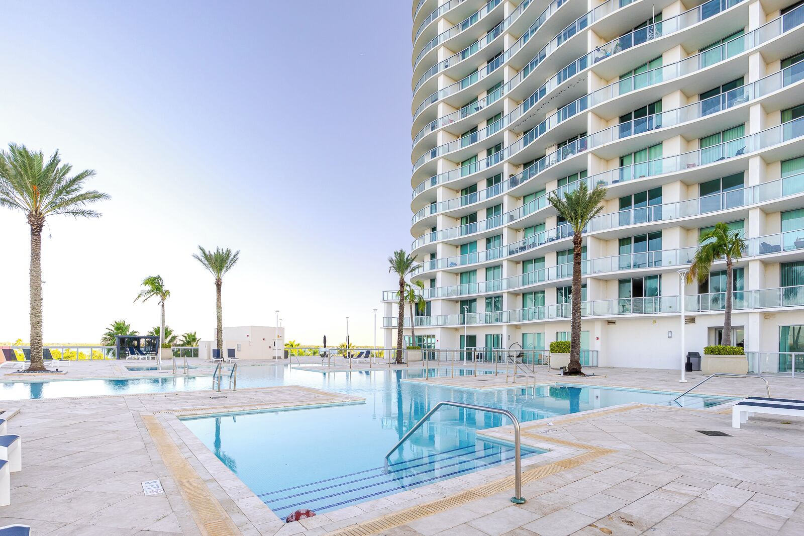 A modern hotel with a large pool, palm trees, and a multi-story building with balconies.