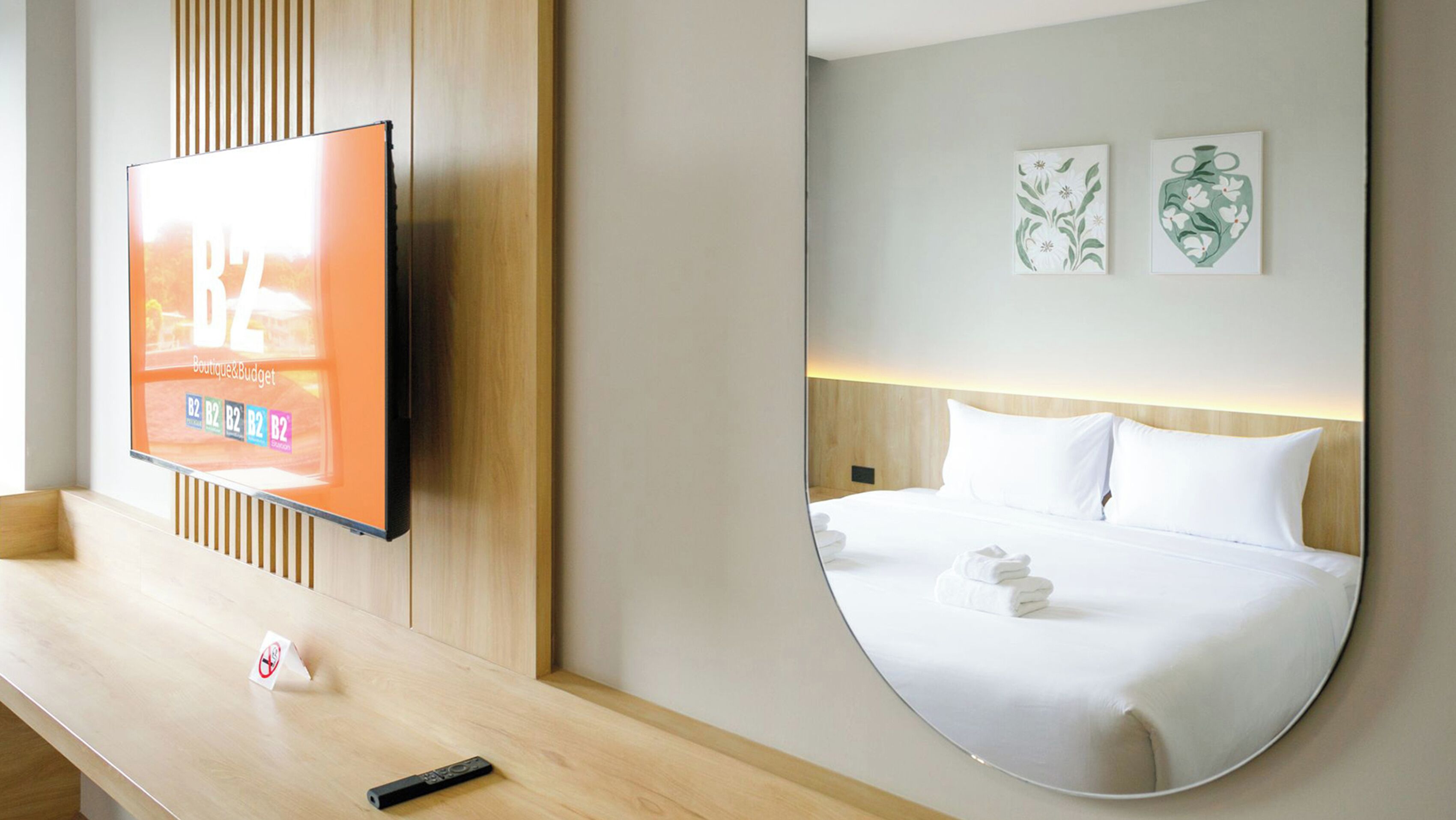 Une chambre d’hôtel dotée d’un grand miroir, d’un lit avec du linge de maison blanc, d’un téléviseur à écran plat fixé au mur et d’un banc en bois avec une télécommande.