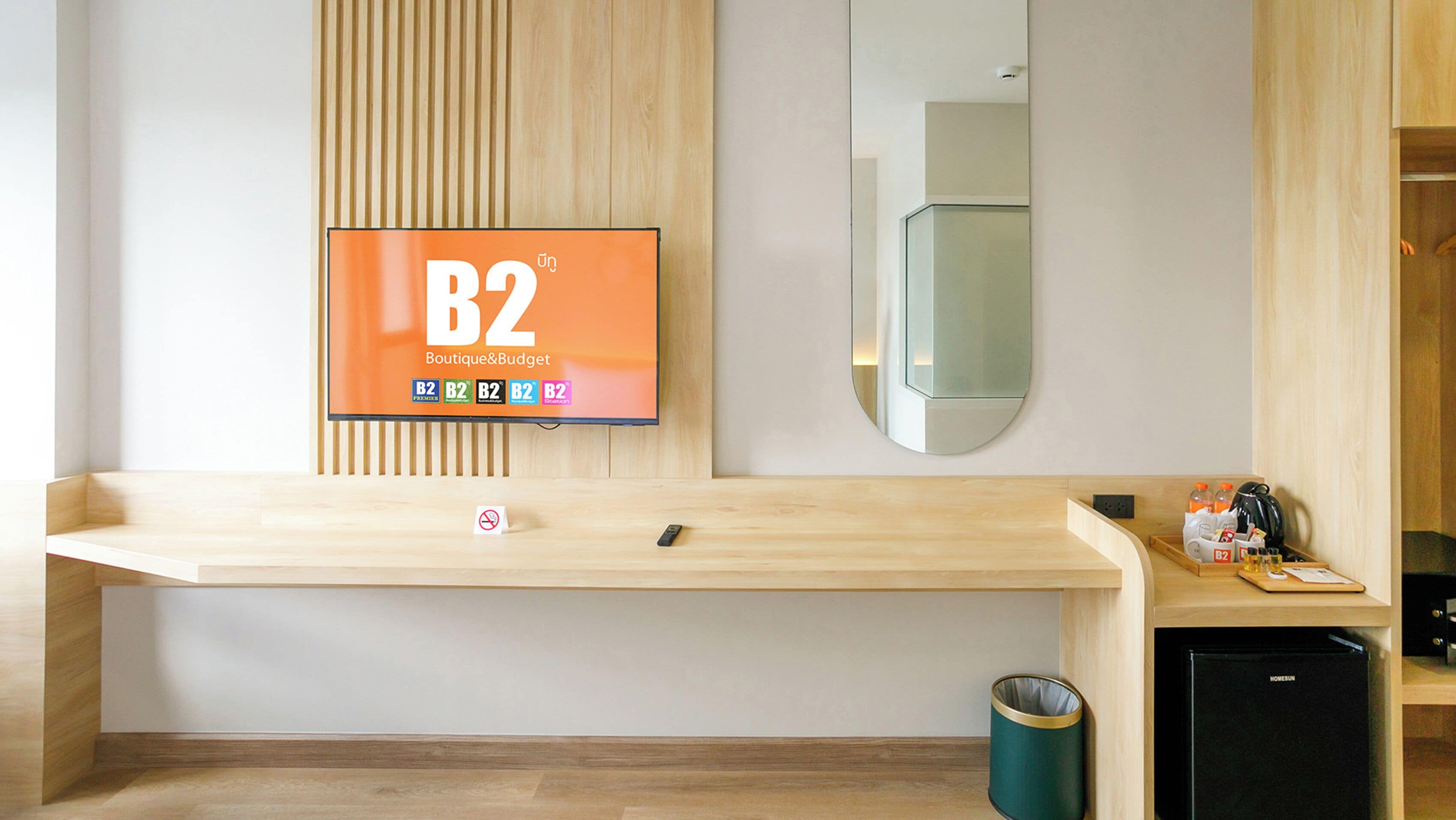A hotel room with a wooden vanity, a B2 Boutique&Budget TV, a mirror, a small trash bin, and a compact refrigerator.