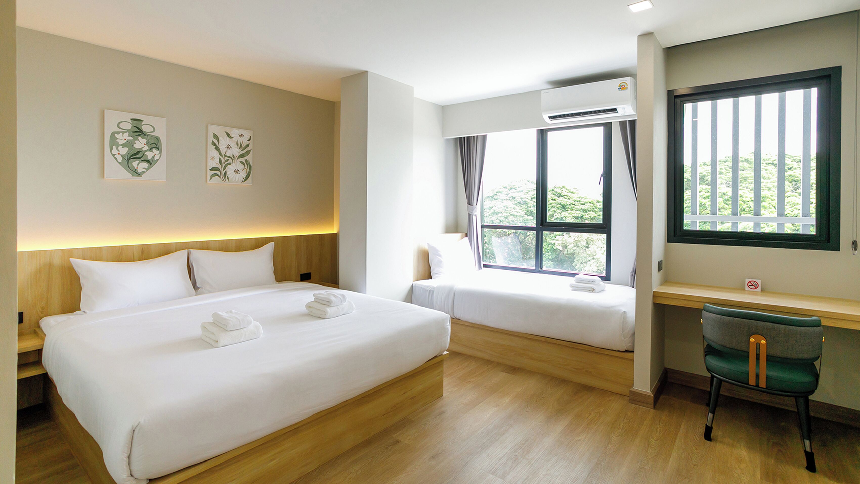 A hotel room with two beds, a desk, and a chair. There are two wall-mounted air conditioning units and a window with curtains.
