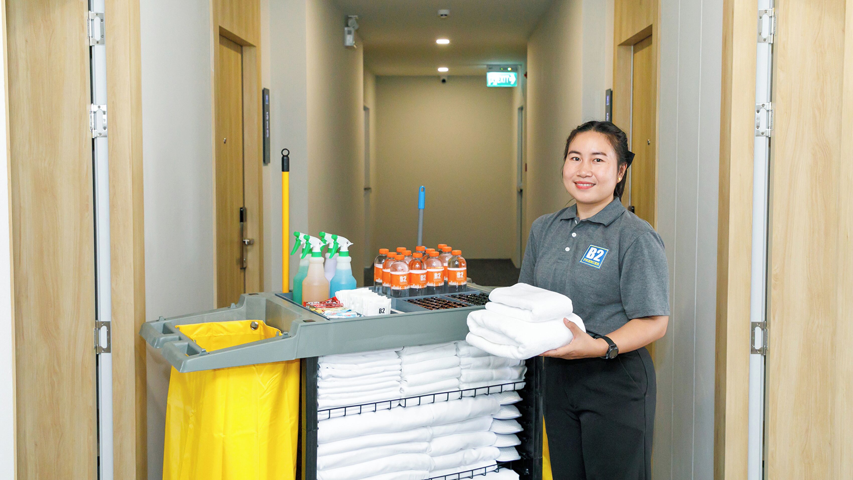 A hotel corridor with a cleaning cart containing cleaning supplies and folded towels.