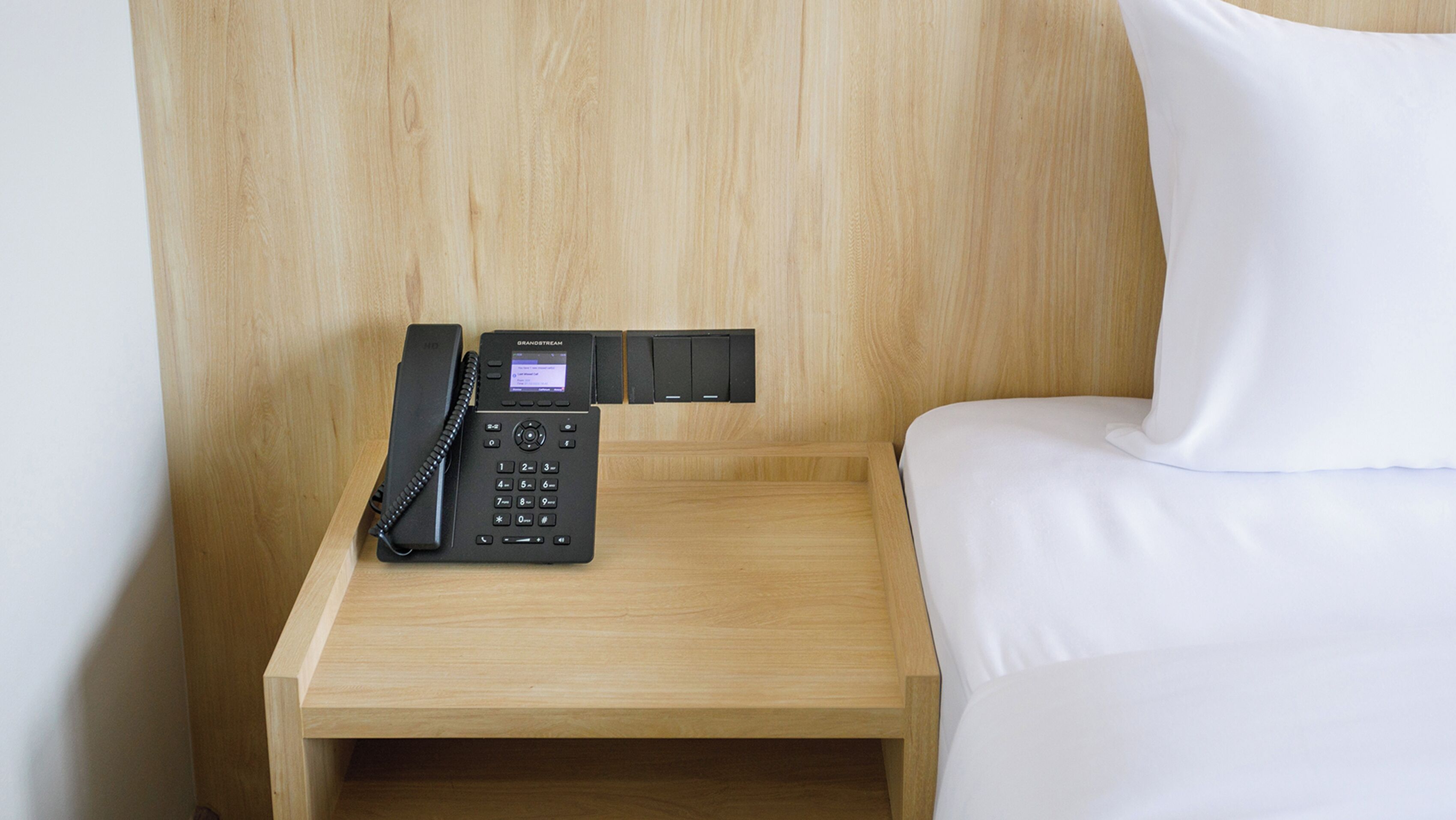 A wooden bedside table with a black telephone and a small control panel.