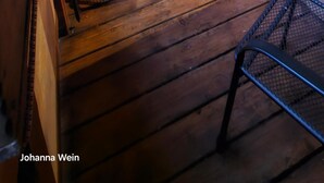 Een houten veranda met een metalen stoel en een kleine tafel, een houten deur met een ruit, en een hanglamp.