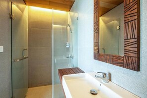 Un baño moderno con ducha envuelta en vidrio, tocador de madera y un espejo grande con marco geométrico.