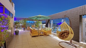 Een modern terras buiten met rieten meubels, een hangstoel en een grote parasol.