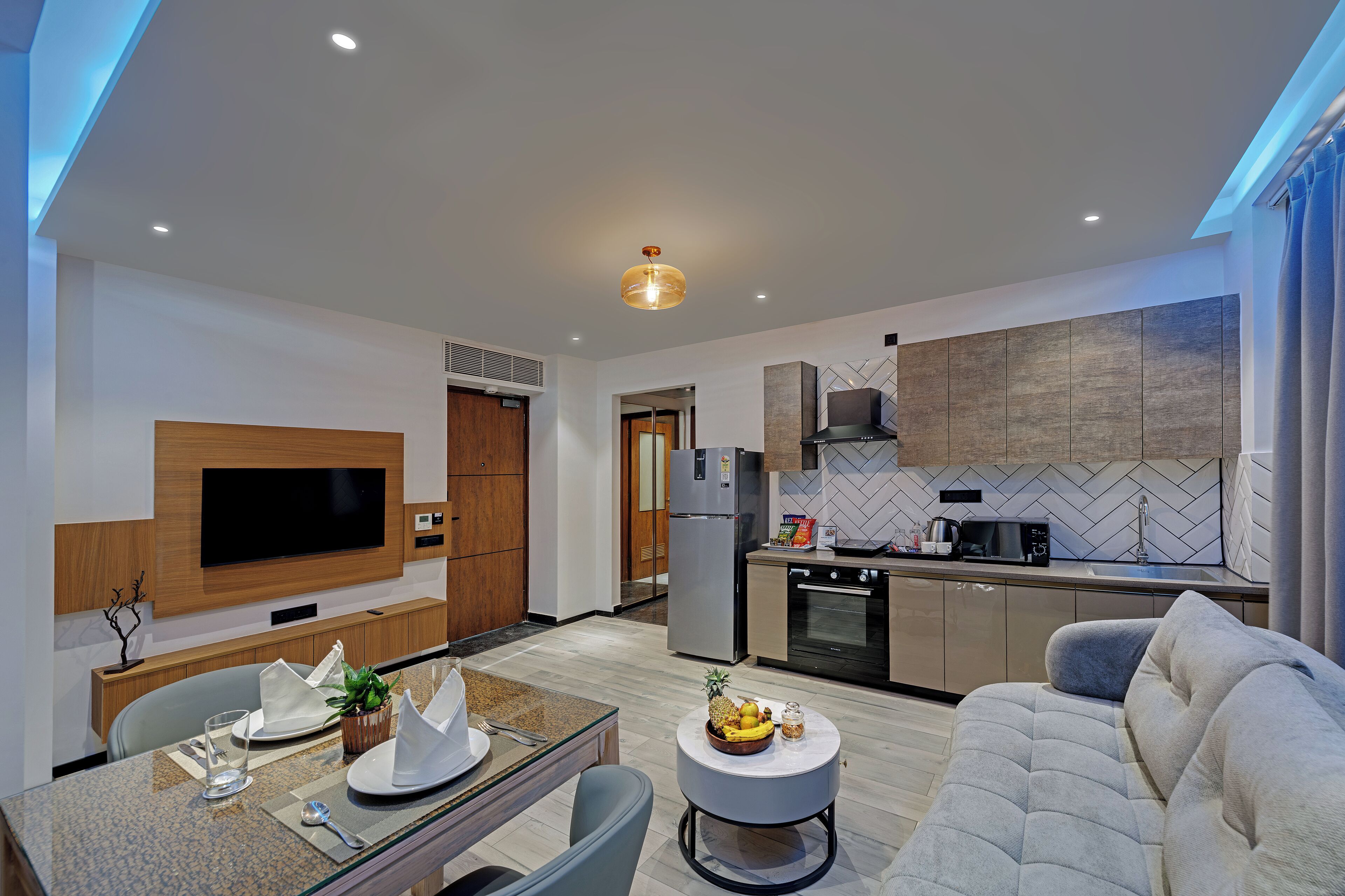 Una cocina moderna con microondas, horno y refrigerador. Hay un comedor con una mesa para dos, un sofá y una pequeña mesa redonda con fruta.