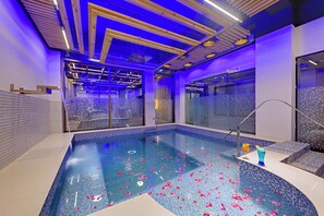 A modern indoor pool with a glass enclosure, blue lighting, and rose petals on the floor.