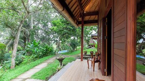 A wooden veranda with a bench, surrounded by lush greenery and a pathway leading to a pool. - Pondok Hari Baik (Selemadeg)