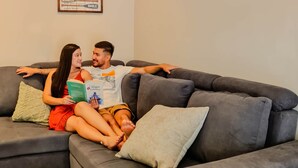 A living room with a grey sofa, a green pillow, and a beige pillow. A poster with the word "Beach" and various words like "ENJOY," "RELAX," and "WALK" is on the wall.