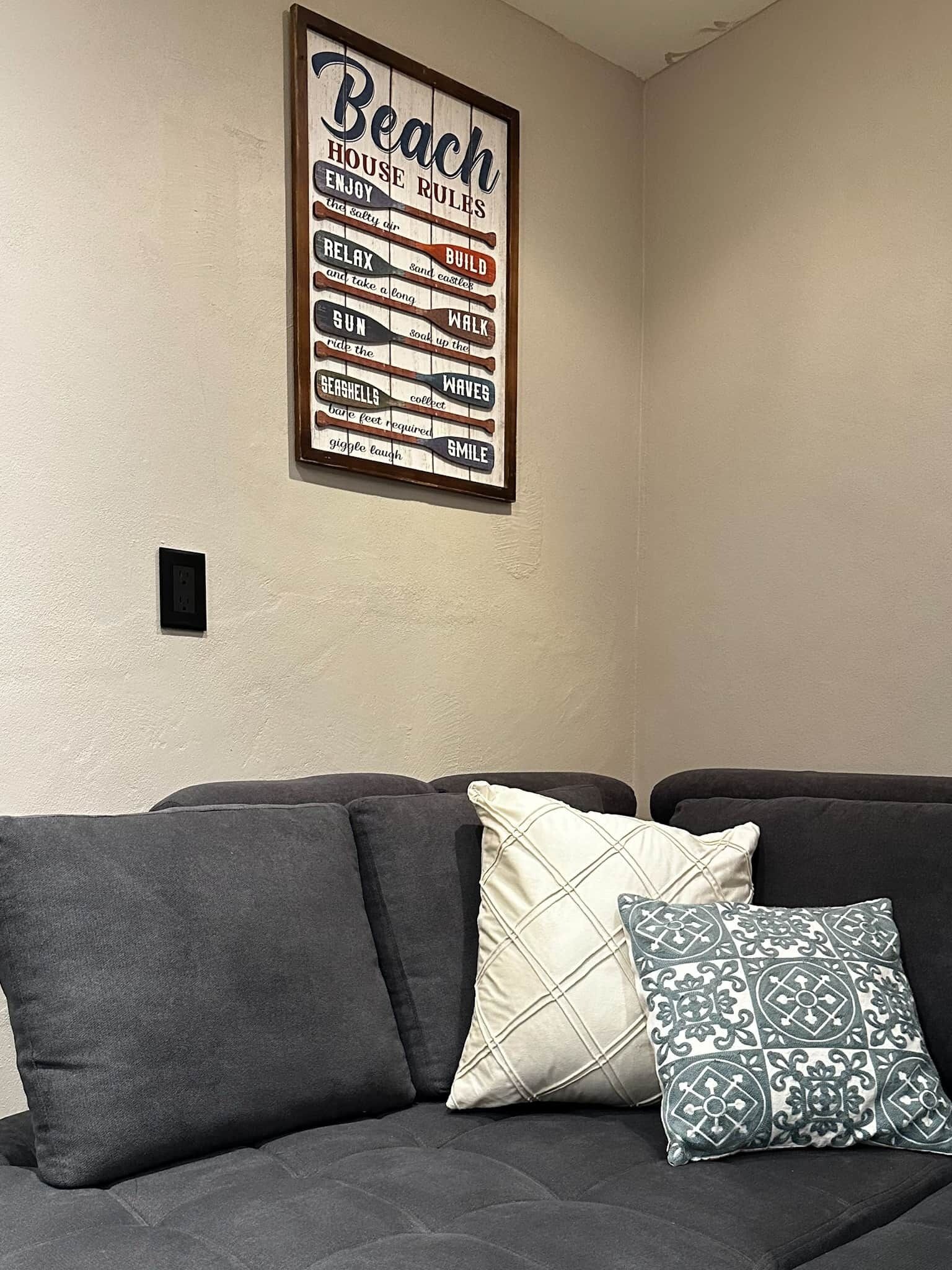 Ein Wohnzimmer mit einem dunkelgrauen Sofa, zwei gemusterten Kissen und einem an der Wand befestigten Schild mit der Aufschrift 'Beach House Rules' und verschiedenen Aktivitäten wie 'ENJOY', 'RELAX', 'BUILD', 'WALK', 'SON', 'SEASHELLS', 'WAVE' und 'SMILE'.