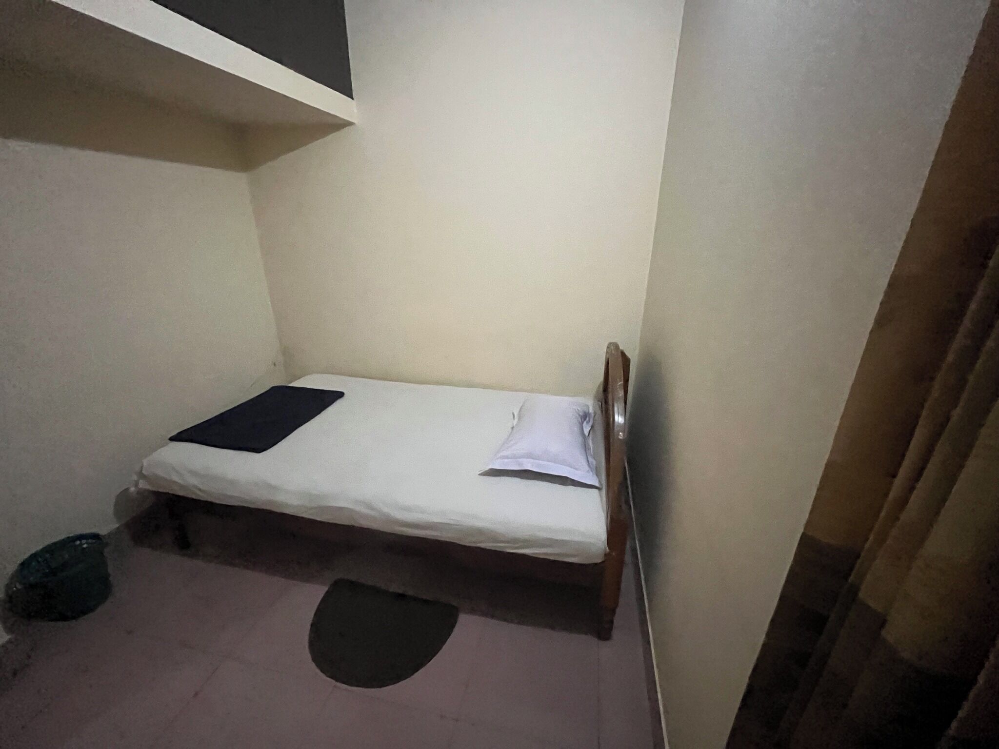 Une petite chambre avec un lit simple, une tête de lit en bois, un matelas blanc et un oreiller noir. Il y a un tapis noir sur le sol et un objet noir sur le côté gauche.