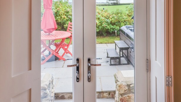 A view through a glass door of an outdoor patio area with a red table and chairs, a pink umbrella, and a barbecue grill. - 1 bedroom accommodation in St Davids (St Davids)