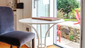 A room with a blue chair, a white table with a book, and a glass door leading to an outdoor area with a red chair and greenery. - 1 bedroom accommodation in St Davids (St Davids)
