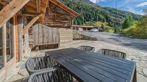 Un edificio rústico de madera con cimientos de piedra, un patio empedrado con mesa y sillas, y una vista de una cadena montañosa al fondo.