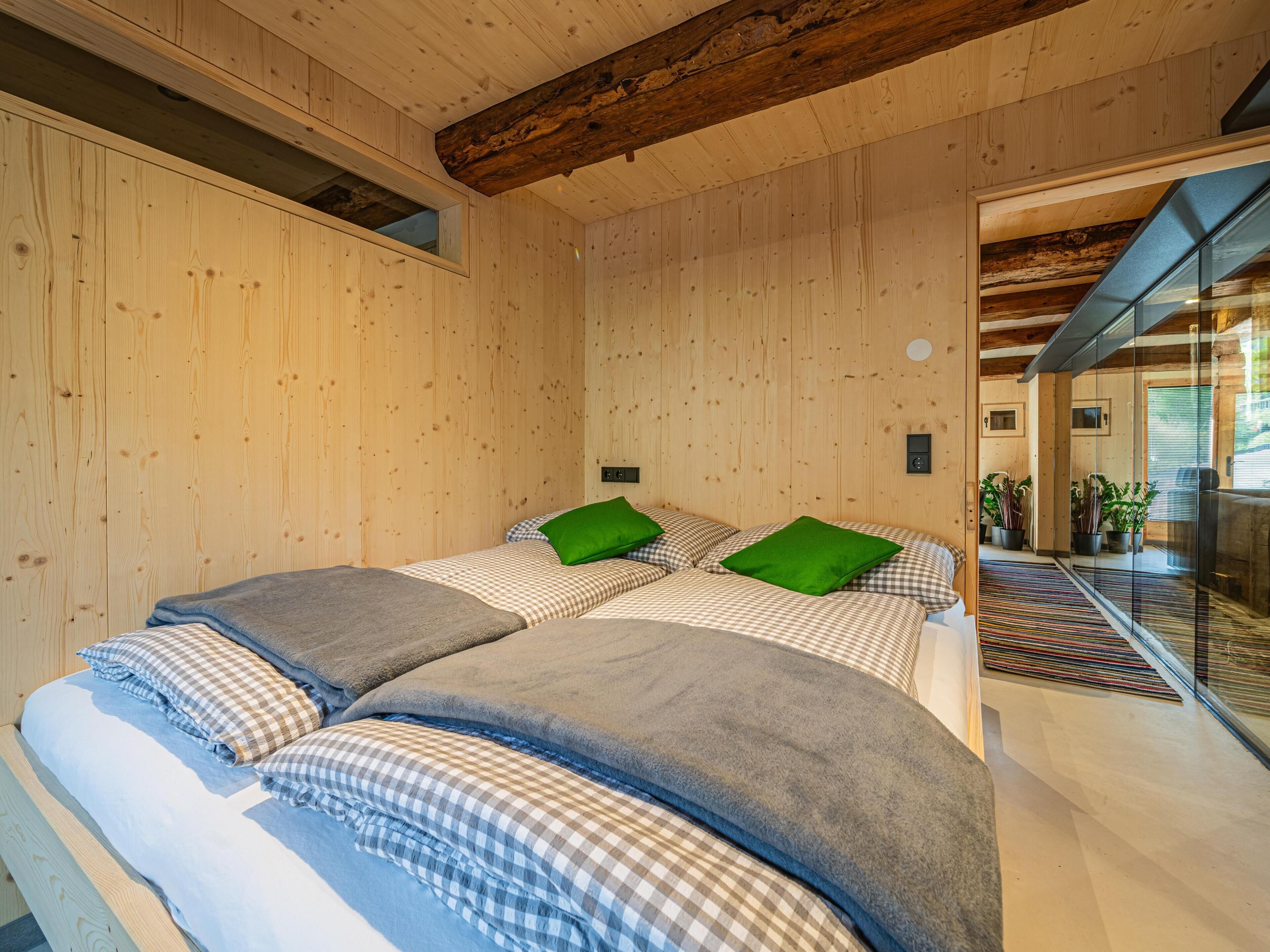 Una habitación de madera con dos camas, ropa de cama a cuadros, almohadas verdes y un ventanal de vidrio que da a un balcón con plantas.