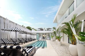 A pool area with lounge chairs and umbrellas, a modern building with large windows, and potted plants. - the den canggu (Canggu)