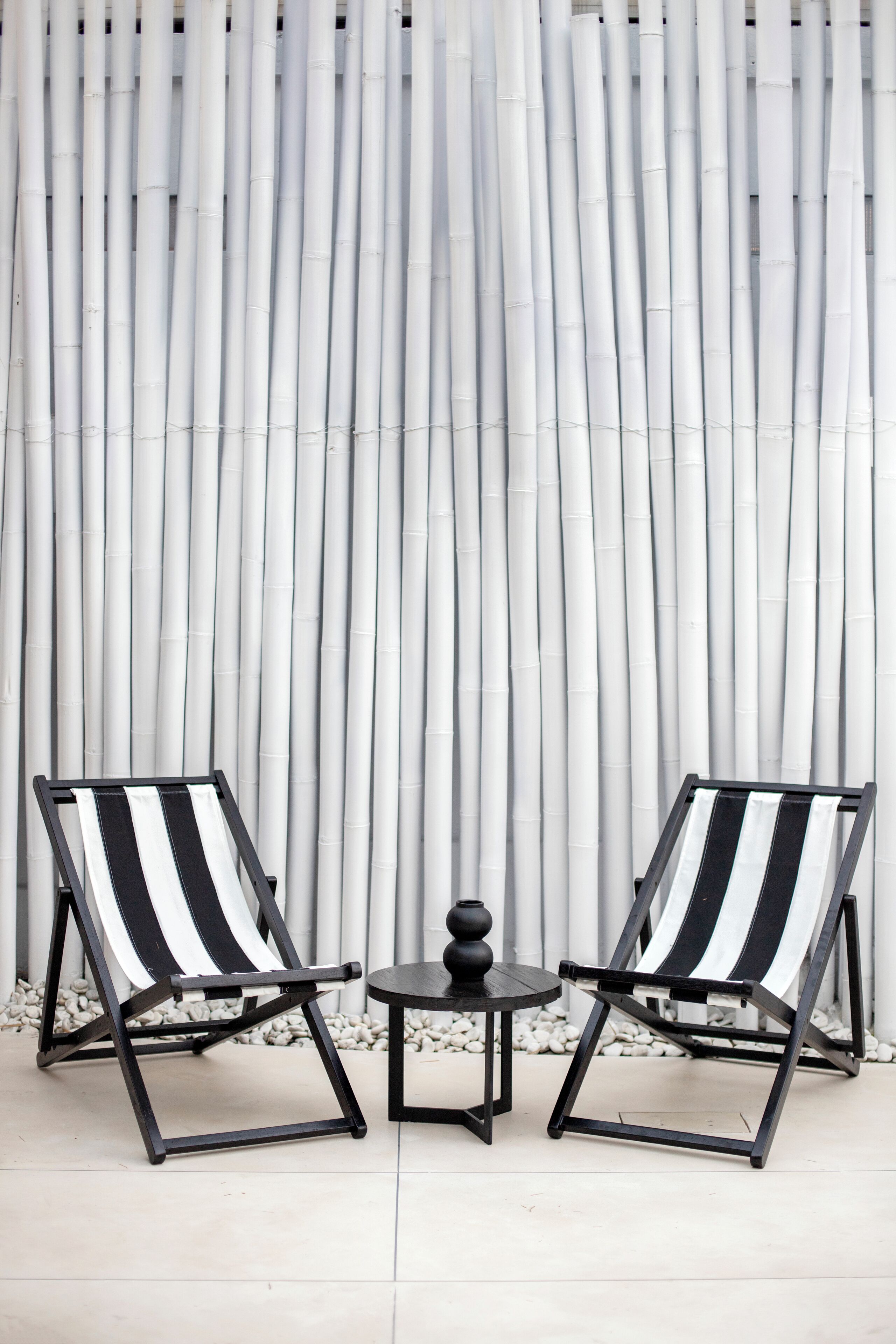 Two black and white striped lounge chairs and a small round table with a vase, set against a backdrop of vertical bamboo panels.