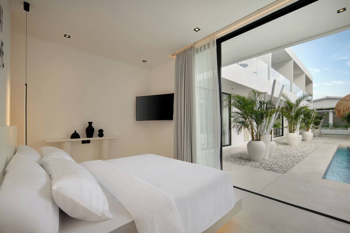 Un dormitorio moderno con una cama grande, un televisor en la pared y vistas a una piscina y plantas al aire libre.