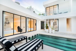 A modern outdoor pool area with lounge chairs, a glass-enclosed room with a bed, and a staircase leading to an upper level. - the den canggu (Canggu)