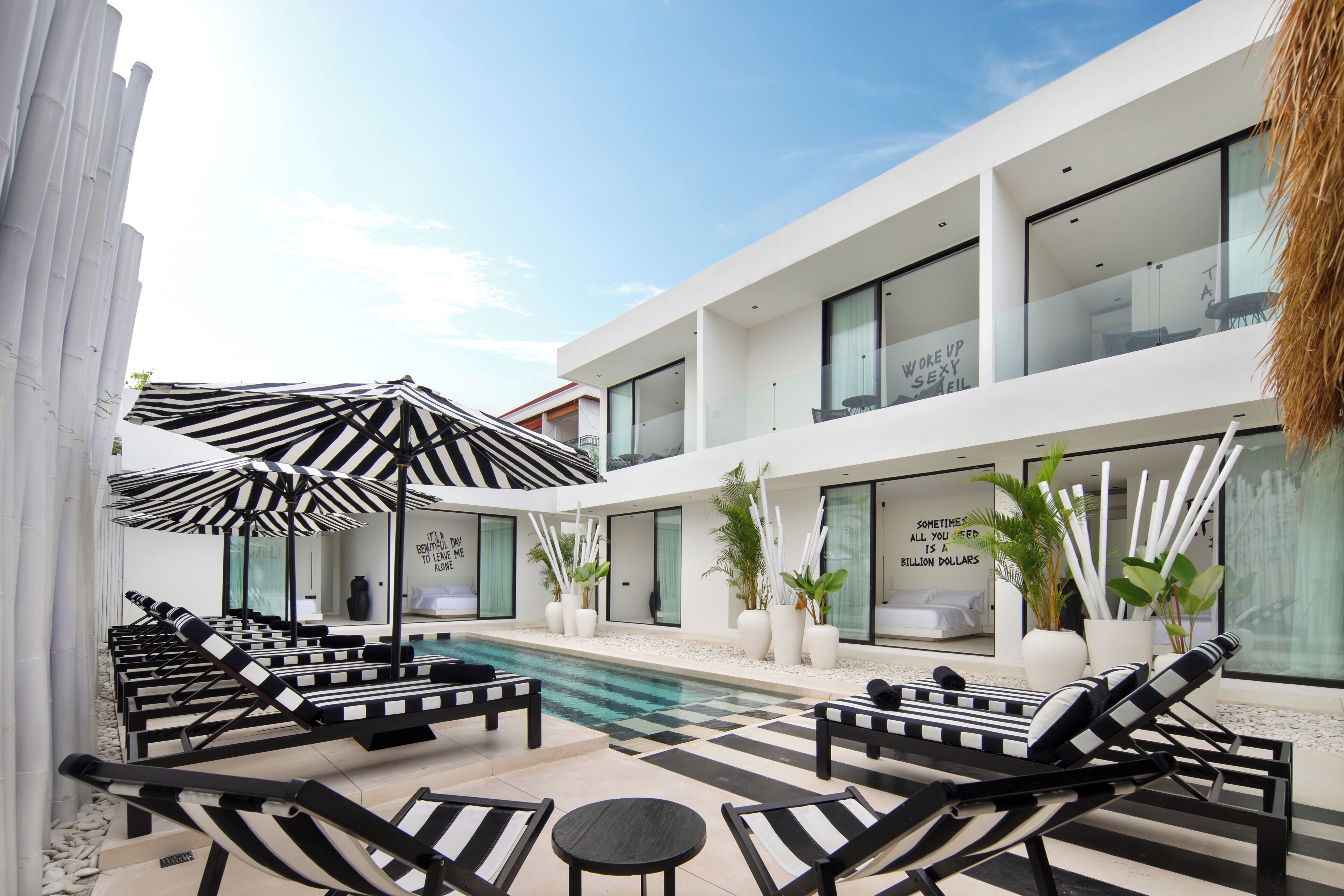 A modern outdoor pool area with lounge chairs and umbrellas, a clear sky, and a building with large windows and a balcony.