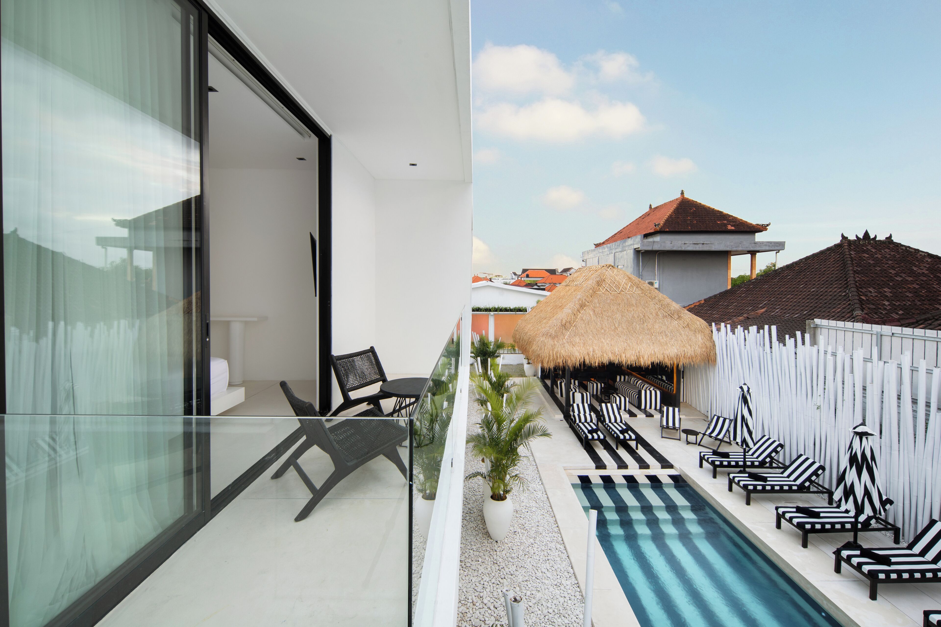 A modern hotel with a glass balcony, a pool area with lounge chairs, and a thatched-roof structure.