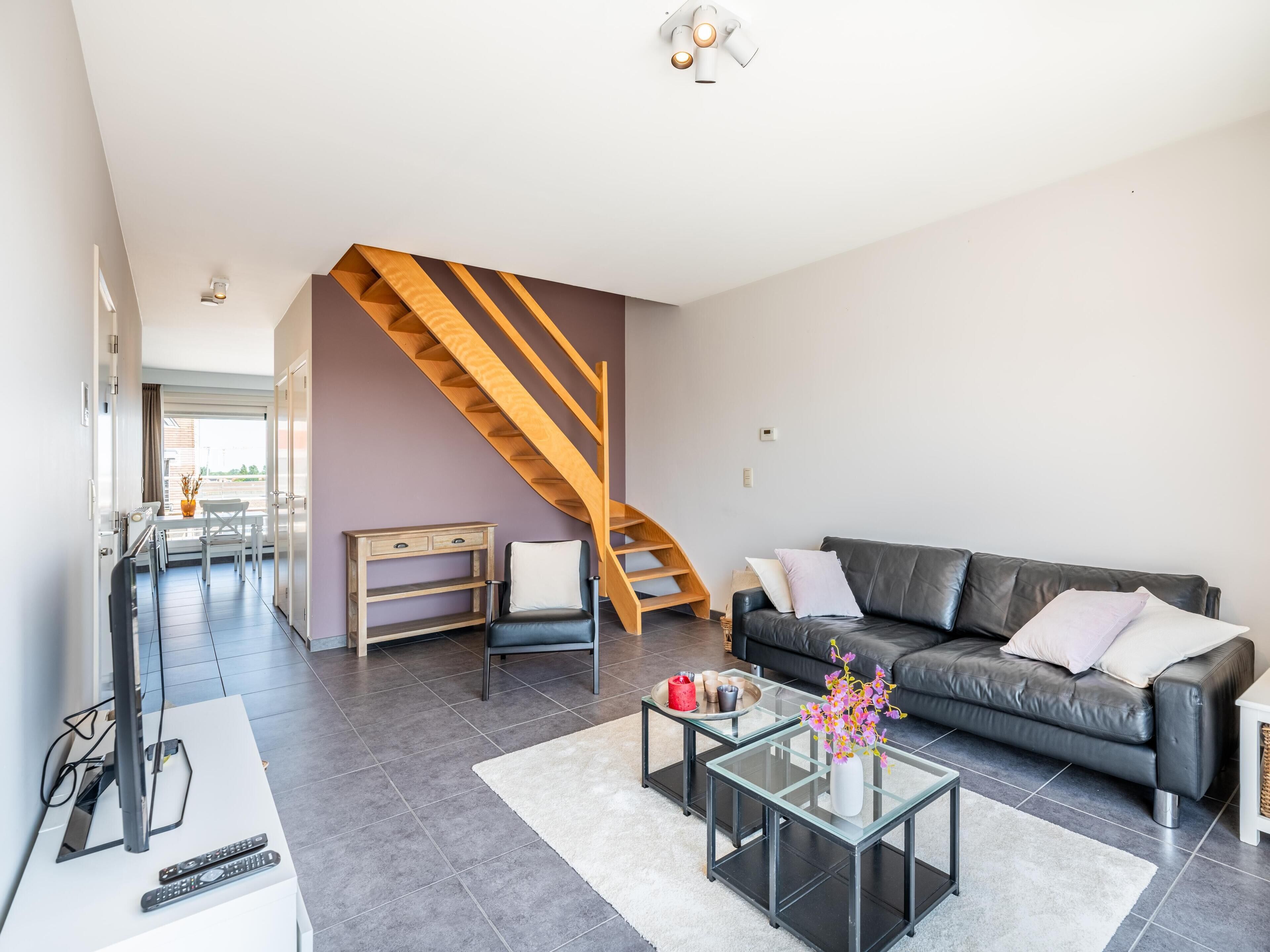 A modern living room with a staircase, a black leather sofa, a glass coffee table, and a white vase with flowers.
