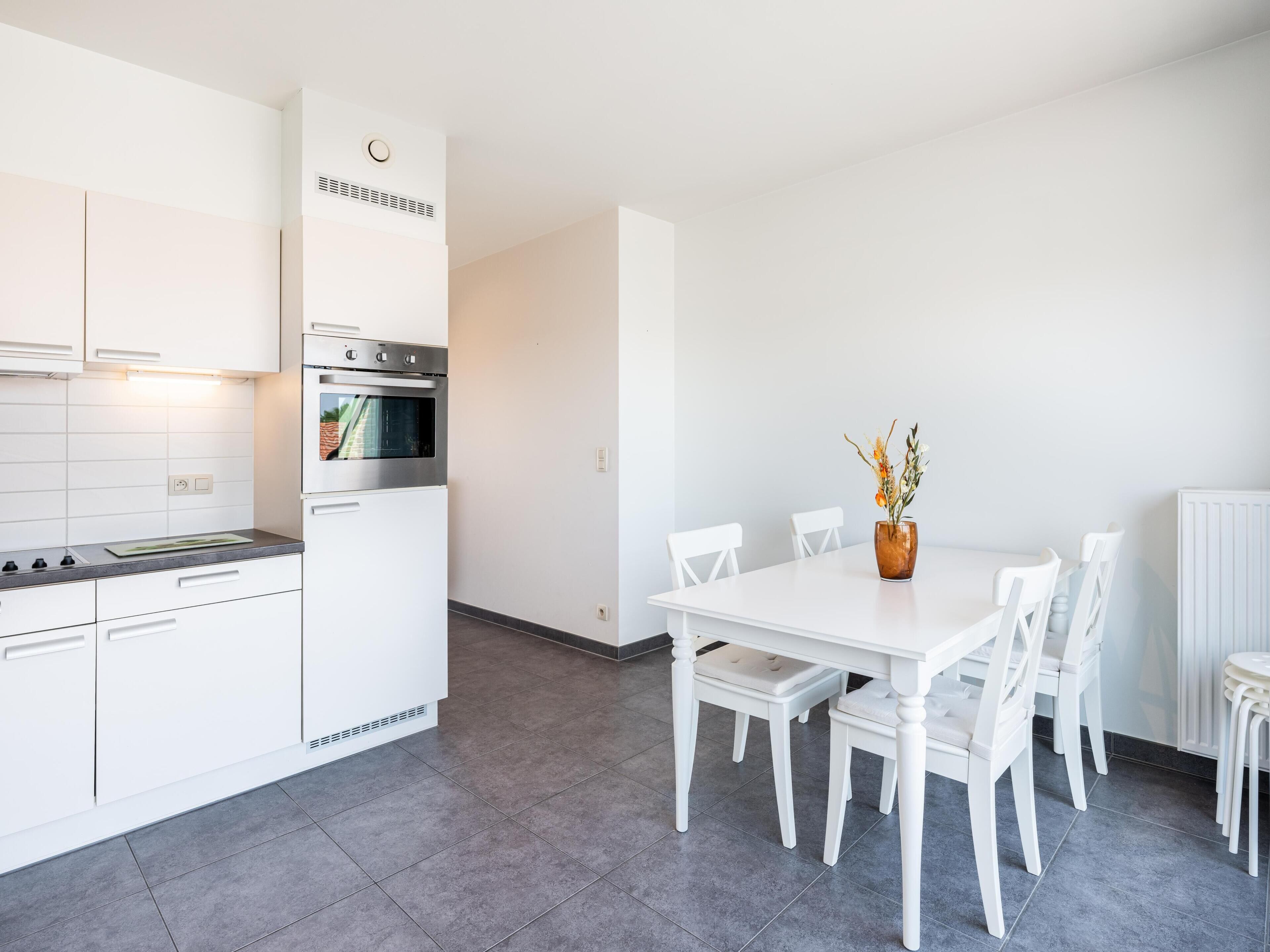 A modern kitchen with white cabinets, a built-in oven, and a dining area with a white table and chairs.