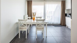 Un appartement moderne doté d’un coin repas avec une table et des chaises blanches, d’une petite cuisine équipée d’un micro-ondes et d’une grande fenêtre agrémentée de rideaux légers.