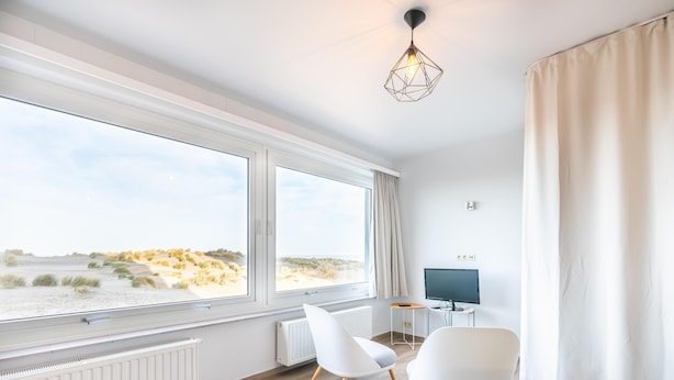 A modern room with large windows, a flat-screen TV, two white chairs, and a geometric pendant light. - Renovated studio with sea views (Oostduinkerke)