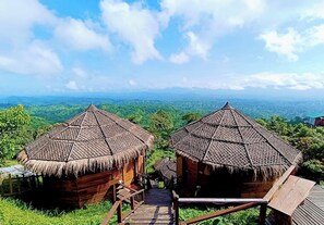 Two thatched-roof huts with wooden staircases leading to them, set against a backdrop of lush green hills and a clear blue sky. - Niribili Eco Resort (Sajek)