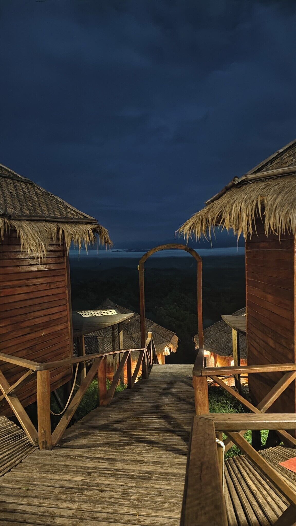 A wooden pathway leading to thatched-roof huts with a view of the landscape.