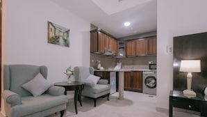 Espaço de estar compacto com sofá, poltrona, mesa pequena e luminária. Uma máquina de lavar e uma área de cozinha são visíveis ao fundo.