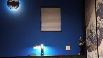Una habitación con una pared azul, una ventana con persianas, un reloj, un detector de humo y una cama con ropa de cama marrón.