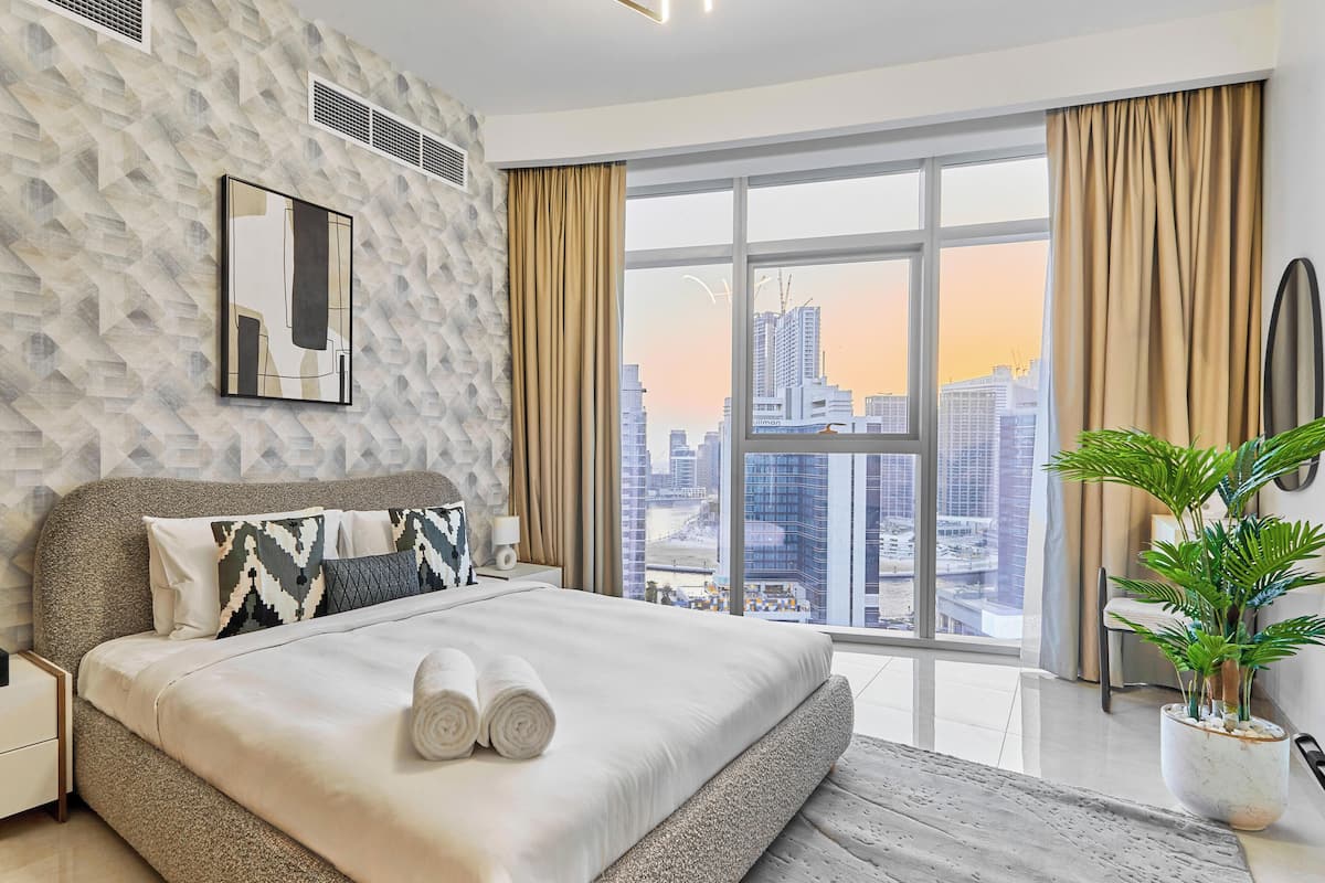 A modern bedroom with a city view, a large bed, and a potted plant.