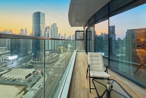 Un immeuble de grande hauteur doté d’un balcon en verre, d’un fauteuil blanc rembourré et offrant une vue sur un paysage urbain avec des gratte-ciel.