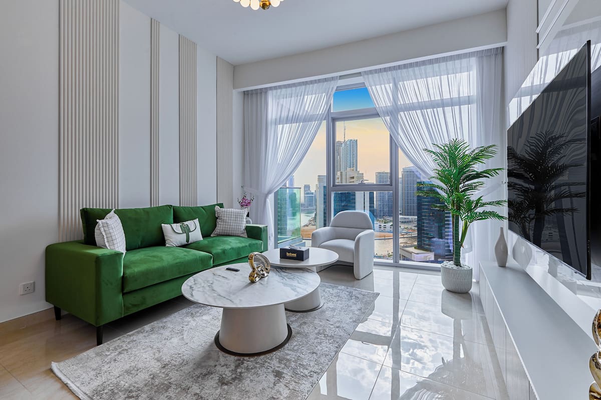 A modern living room with a green sofa, a white coffee table, and a large window offering a city view.