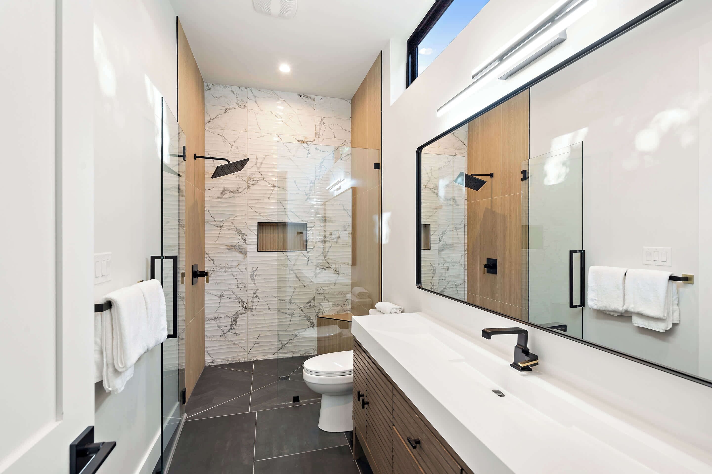 A modern bathroom with a glass shower enclosure, a marble backsplash, a white toilet, and a large mirror above the sink.