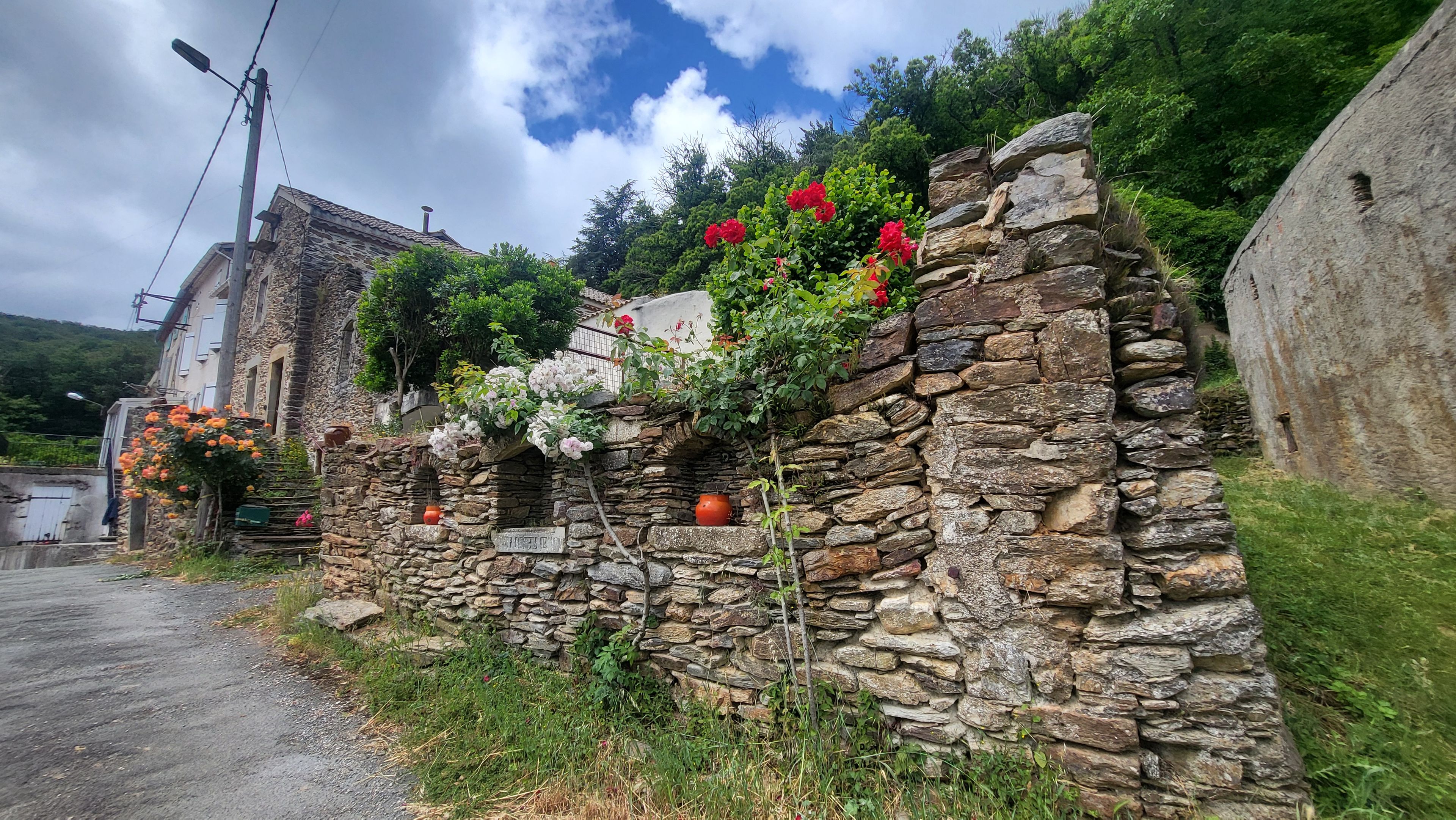 A stone wall with a rustic stone archway, a white building with a sloped roof, and a lush green hillside in the background.