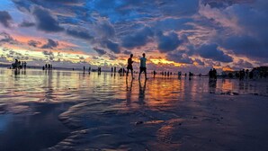 A beach scene with people walking and standing, reflecting on the wet sand under a colorful sunset sky. - Hotel Opera Ocean (Cox's Bazar)