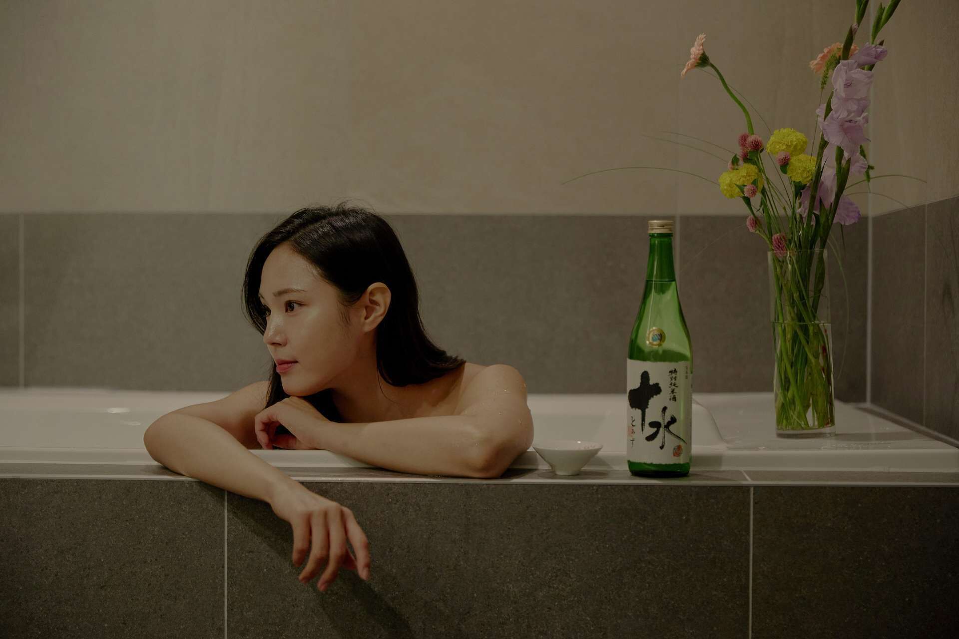 A person in a bathtub with a bottle and a vase of flowers on the edge.