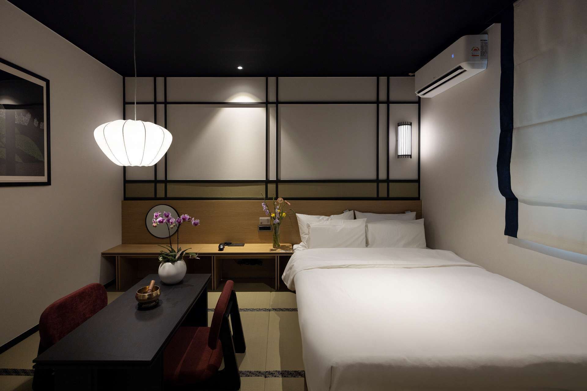 Una habitación de hotel con cama, mesita de noche, escritorio, silla y una lámpara colgante.