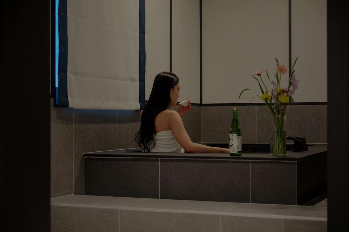 A woman in a white towel sitting by a bathtub with a bottle and a vase of flowers on the counter.