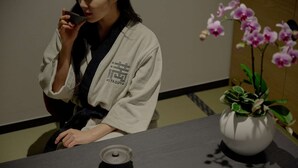 A person in a white robe sitting at a table with a black speaker and a white vase with pink orchids. - Mitasuya Ryokan Hotel by Anook Daejeon Yuseong (Daejeon)