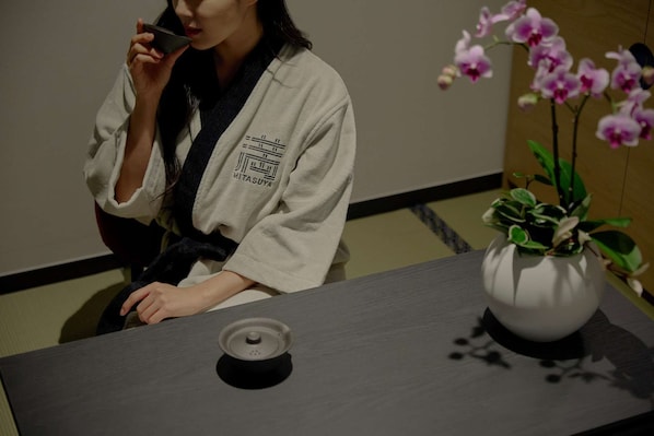 A person in a white robe sitting at a table with a black speaker and a white vase with pink orchids. - Mitasuya Ryokan Hotel by Anook Daejeon Yuseong (Daejeon)