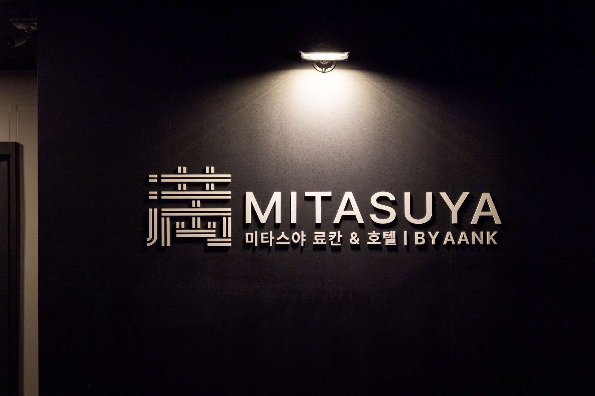 A wall with the name 'MITASUYA' and the text 'i'taaswa yookan & hoetil byaank' in Korean, illuminated by a spotlight.