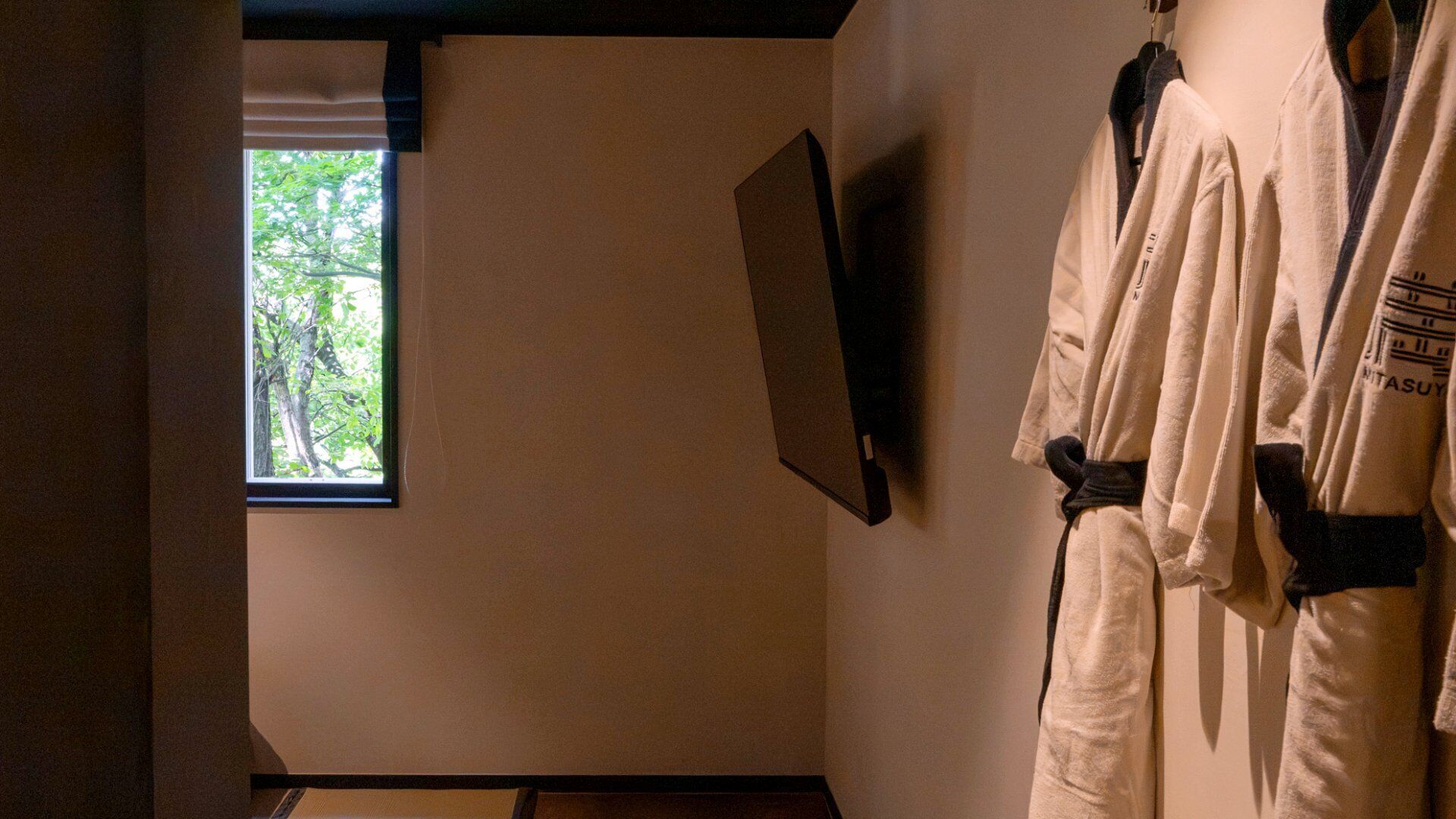 A room with a window, a flat-screen TV mounted on the wall, and a white robe hanging on a hook.