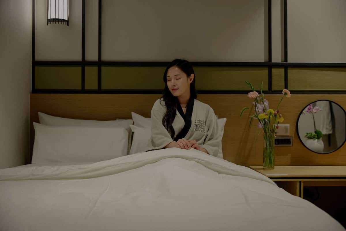 A hotel room with a bed, pillows, a bedside table with a vase of flowers, and a mirror on the wall.