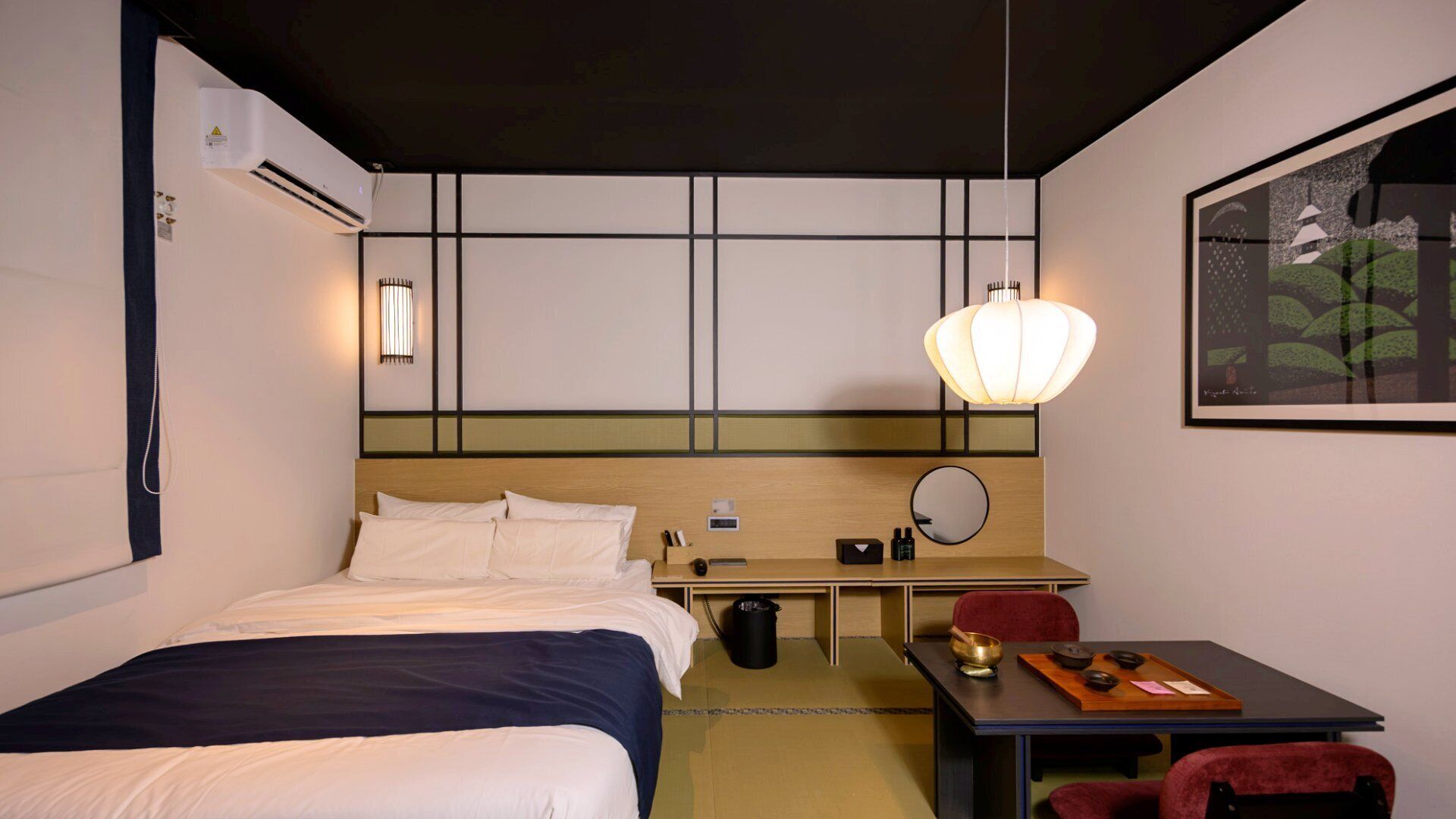 A hotel room with a bed, bedside table, desk, chair, and a painting on the wall.