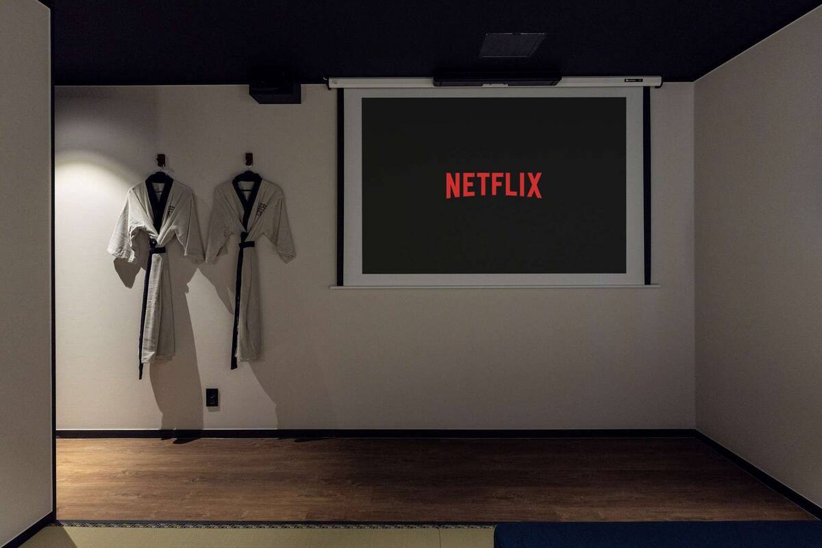 A room with a projector screen displaying "NETFLIX" in red, two white robes hanging on the wall, and a wooden floor.
