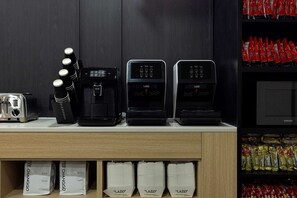 Un coin cuisine équipé d’un grille-pain, d’une machine à café et de deux machines à café LAZO. On y trouve également des piles de gobelets et de dosettes de café jetables, rangées sur des étagères.